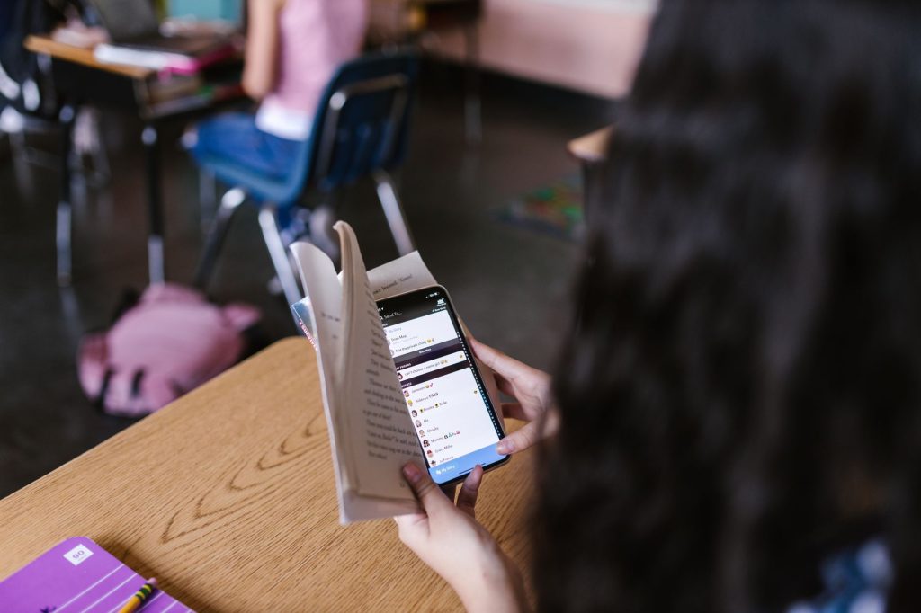 A student in a classroom multitasks by reading a book while using a smartphone, emphasizing modern educational challenges. 文章配图