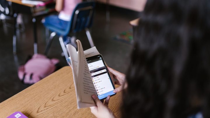 A student in a classroom multitasks by reading a book while using a smartphone, emphasizing modern educational challenges. 文章配图