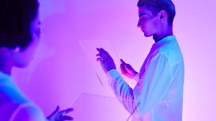Two individuals engaging with futuristic transparent touch screens in a vibrant purple-lit studio.