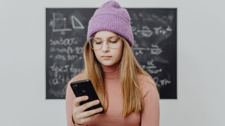 Teenager wearing a beanie and eyeglasses using a smartphone against a blackboard.